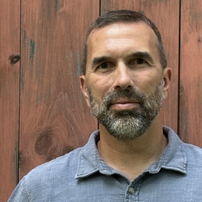 A man with short brown hair and a gray beard, wearing a light blue button-up shirt, stands in front of a wooden fence with a serious expression, as if preparing for Operation Outbreak.