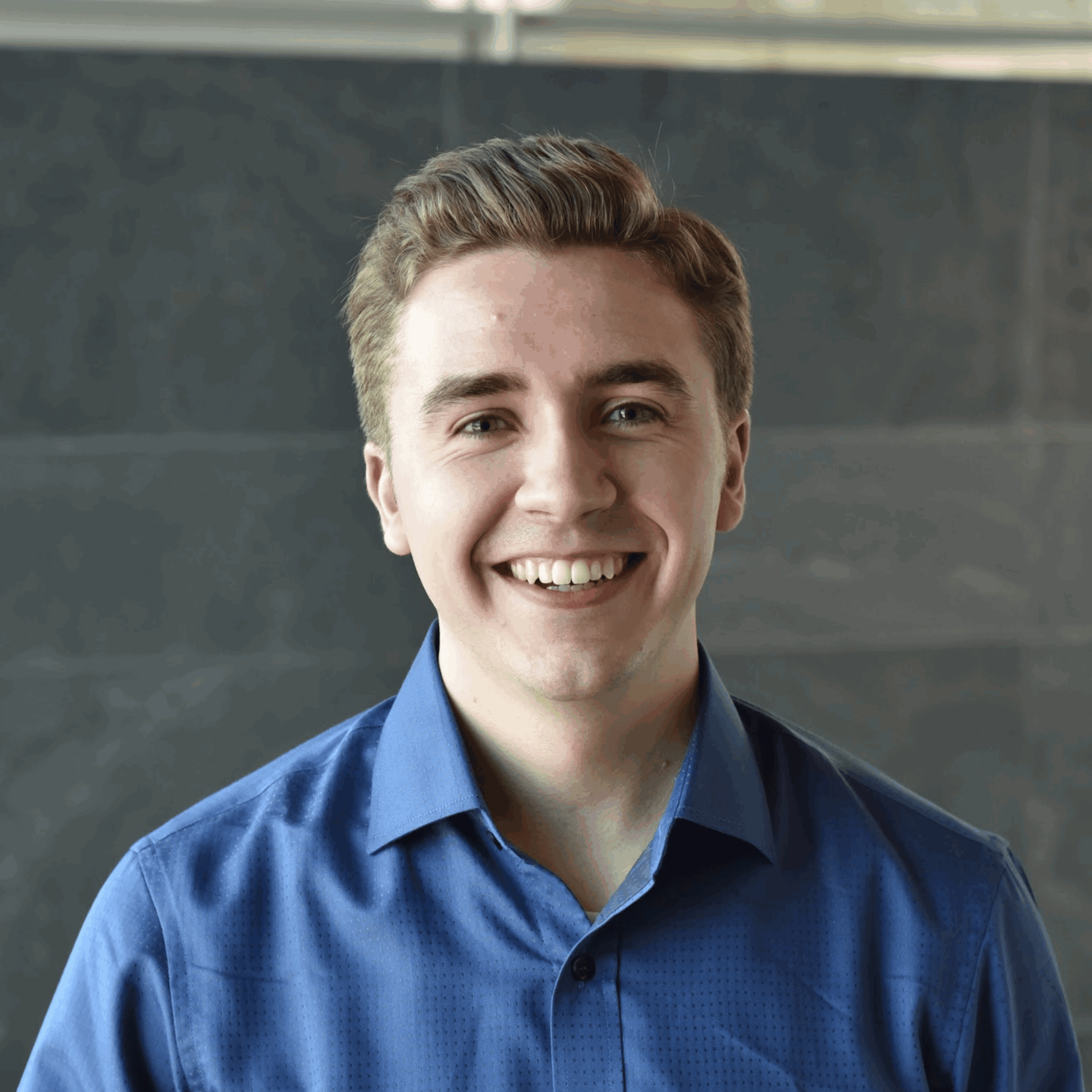 A young man with short light brown hair smiles at the camera. He is wearing a blue button-up shirt and standing in front of a dark, blurred background.