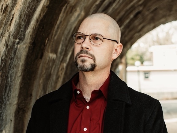 A bald man with glasses, a goatee, and a mustache wearing a red shirt and black coat stands under a stone archway, looking thoughtfully into the distance.