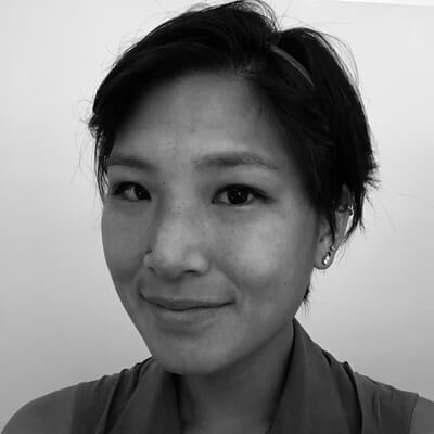 A person with short dark hair, wearing a sleeveless top and small hoop earrings, smiles softly at the camera in a black-and-white portrait against a plain background, evoking the candid spirit of Operation Outbreak.