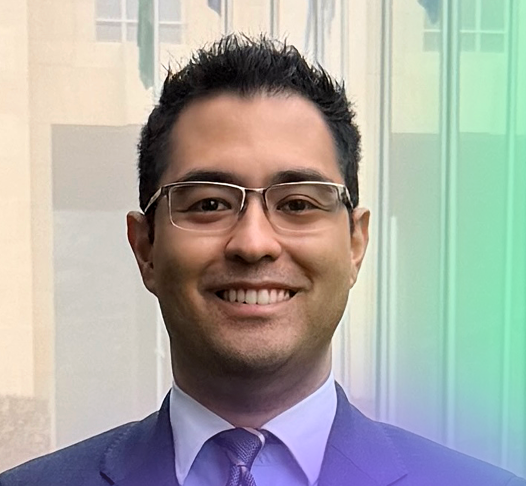 A man with short dark hair and glasses, wearing a suit and tie, smiles at the camera in front of a glass building with faint reflections and a gradient overlay at the bottom.