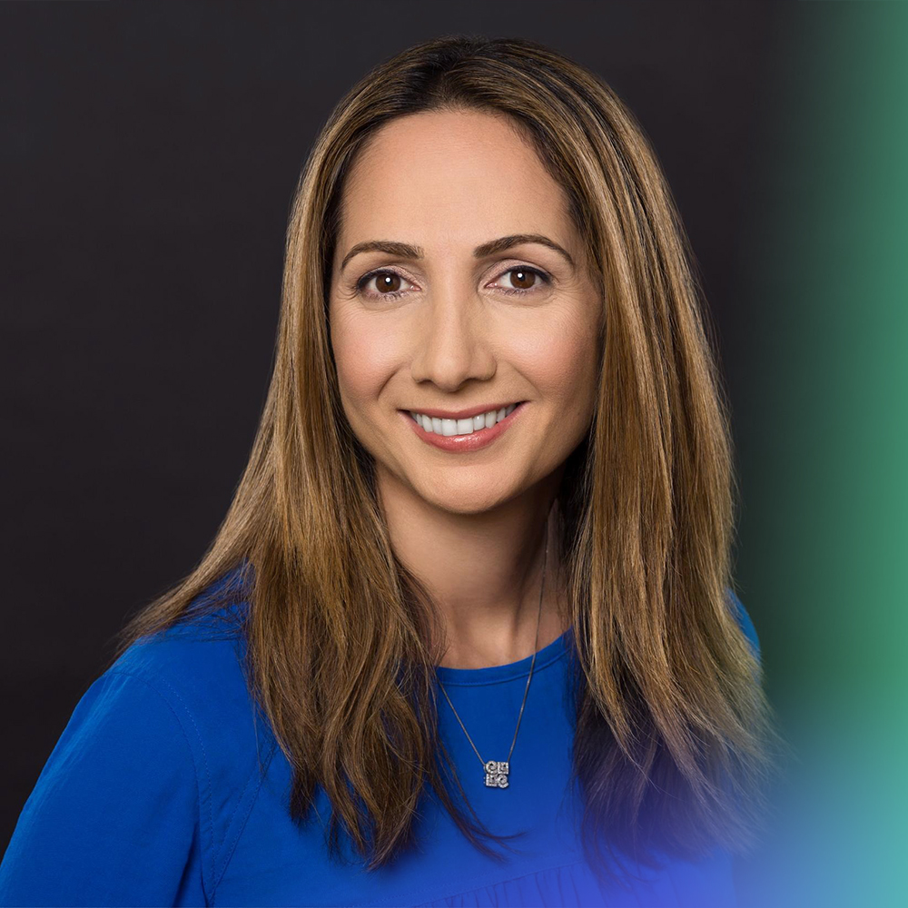 A woman with long, straight brown hair smiles at the camera. She is wearing a blue top and a necklace, with a dark background and a blurred green gradient on the right side of the image.