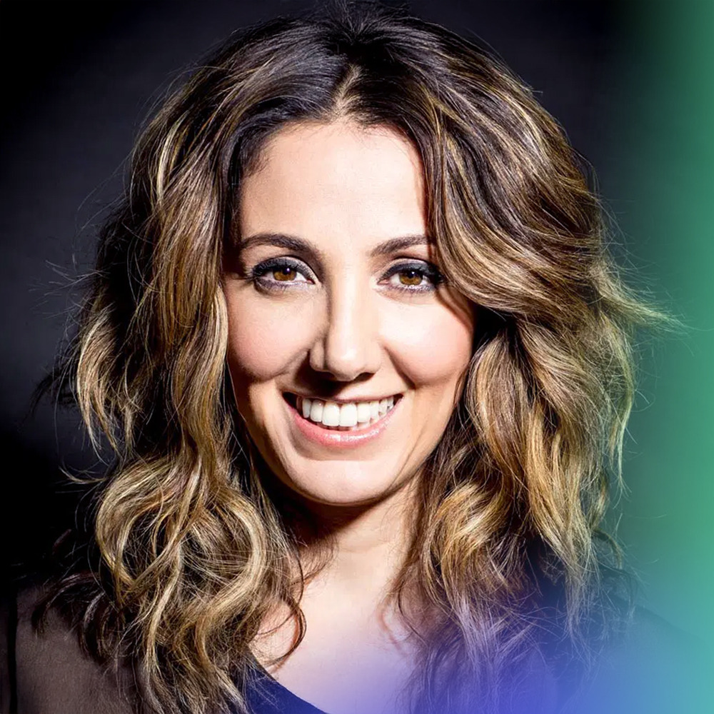 A smiling woman with wavy, highlighted brown hair, wearing a dark top, poses against a dark background with a soft green and blue gradient in the lower right corner.