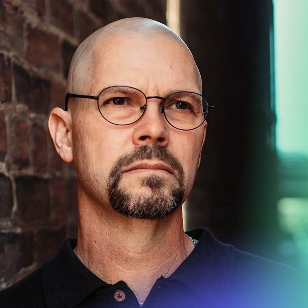 A middle-aged man with a bald head, trimmed goatee, and glasses stands in front of a brick wall, looking thoughtfully into the distance. He is wearing a black collared shirt.