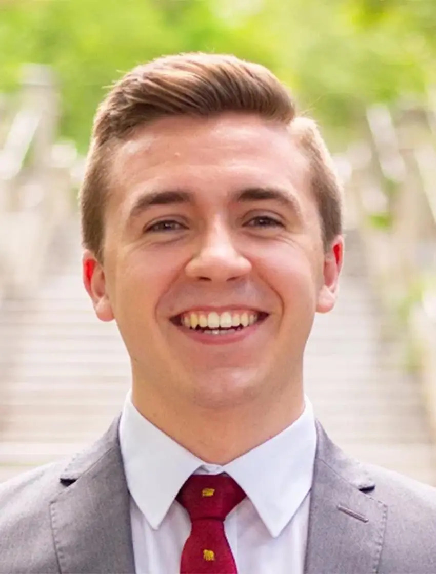 A young man in a grey suit and red tie smiles outdoors, with blurred green trees and stairs in the background.