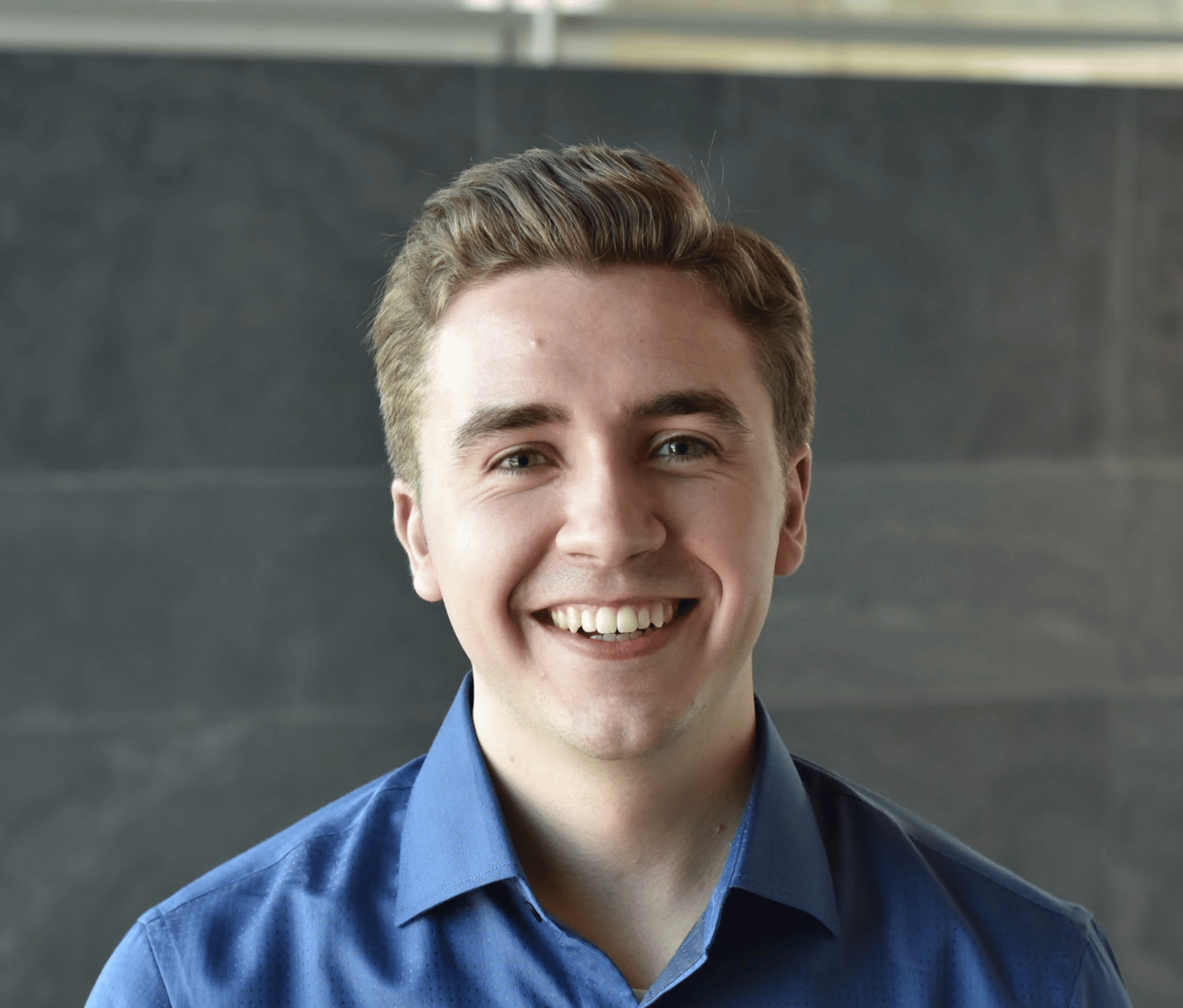 A young man with light brown hair, wearing a blue collared shirt, smiling at the camera. The background is a plain, dark gray wall.