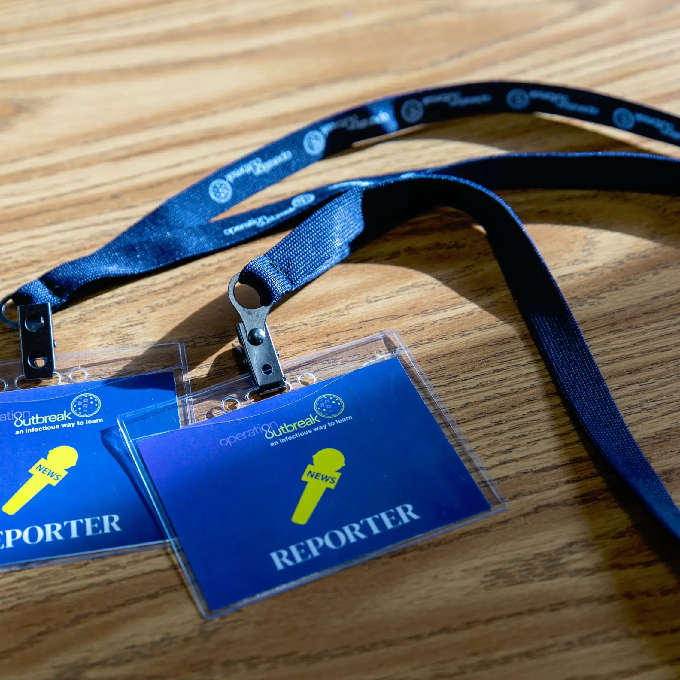 Two blue lanyards with clear plastic badge holders labeled “REPORTER” and a yellow “NEWS” icon lie on a wooden surface, catching sunlight—a scene from Operation Outbreak coverage—casting dynamic shadows.