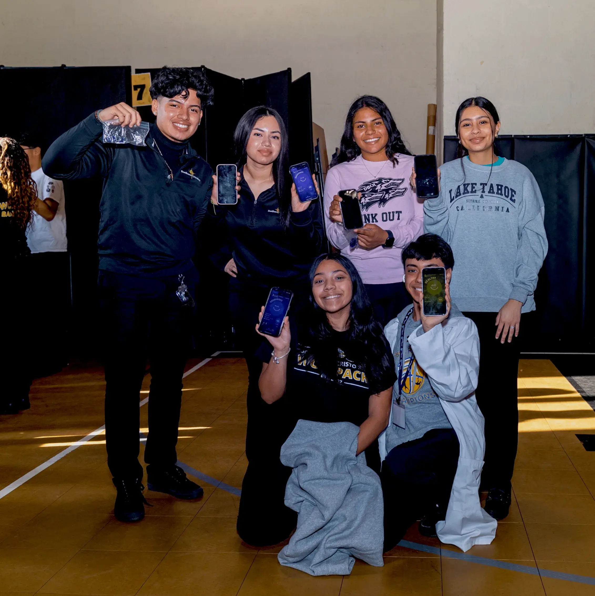 Six smiling young people pose indoors, holding up their phones toward the camera during Operation Outbreak. Five are standing, one is kneeling in front. They wear casual clothes, with black curtains and a gym floor visible in the background.