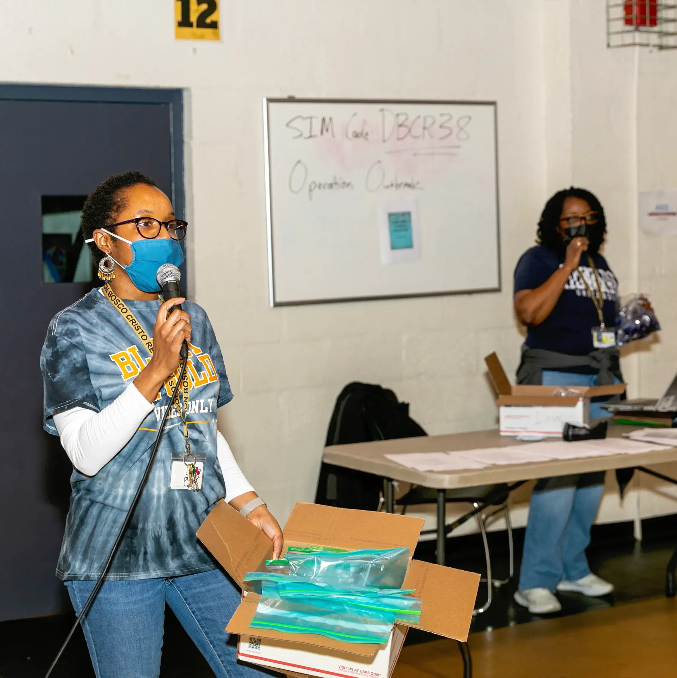 Two women wearing masks stand in a room during Operation Outbreak; one holds a microphone and a box with plastic bags, while the other stands by a table with supplies. A whiteboard with writing is visible in the background.