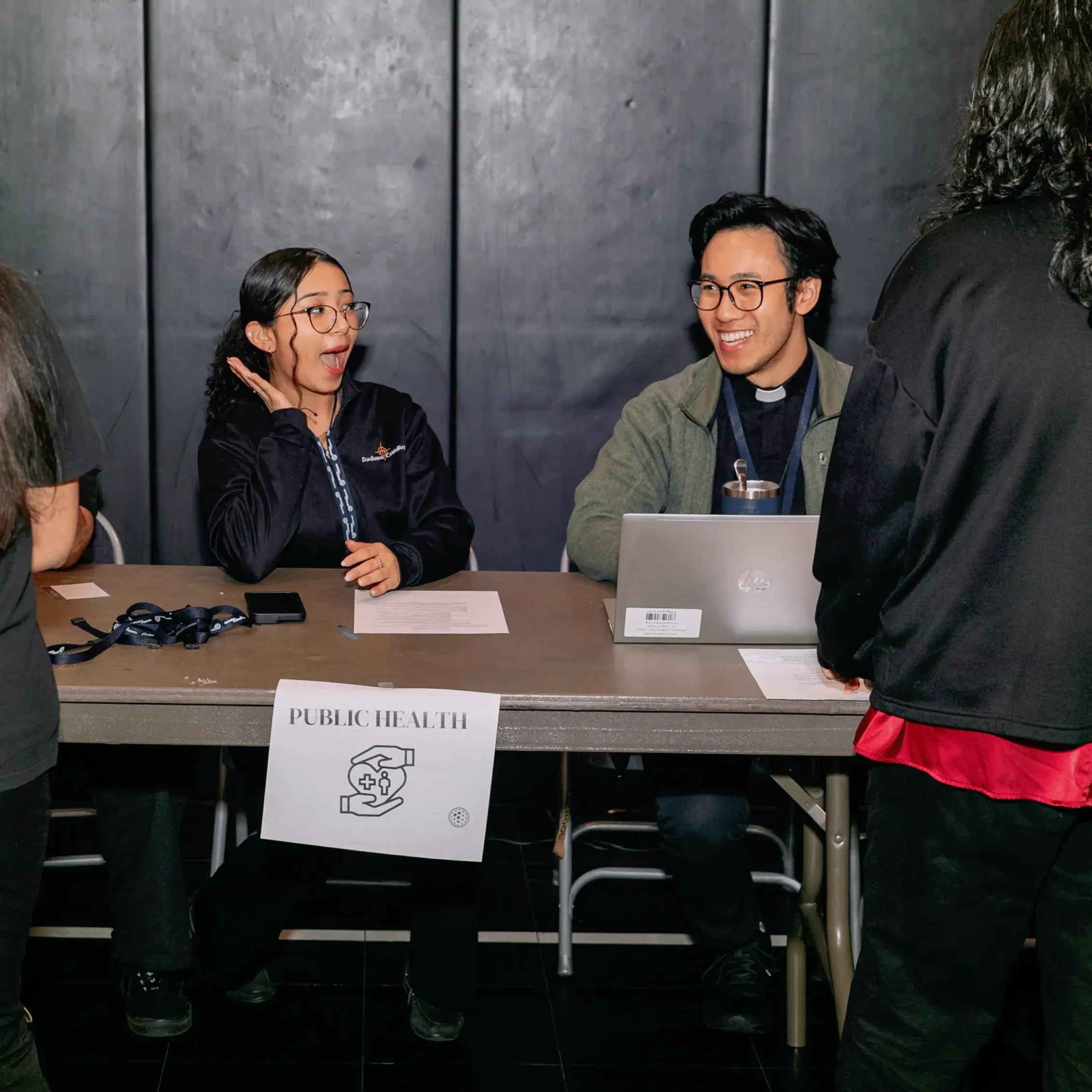 Two people sit behind a table with an “Operation Outbreak” Public Health sign, papers, and a laptop, talking to others who are standing in front of the table at the event. One seated person is smiling with a surprised expression.