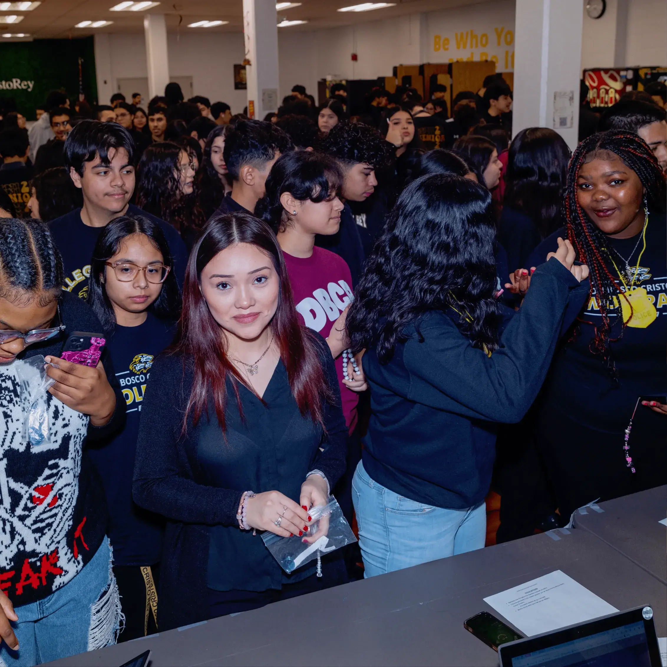 A diverse group of high school students stands in a crowded room during Operation Outbreak, some smiling and talking. A girl in the front with straight brown hair looks at the camera while holding a small bag, with more students and school decor in the background.
