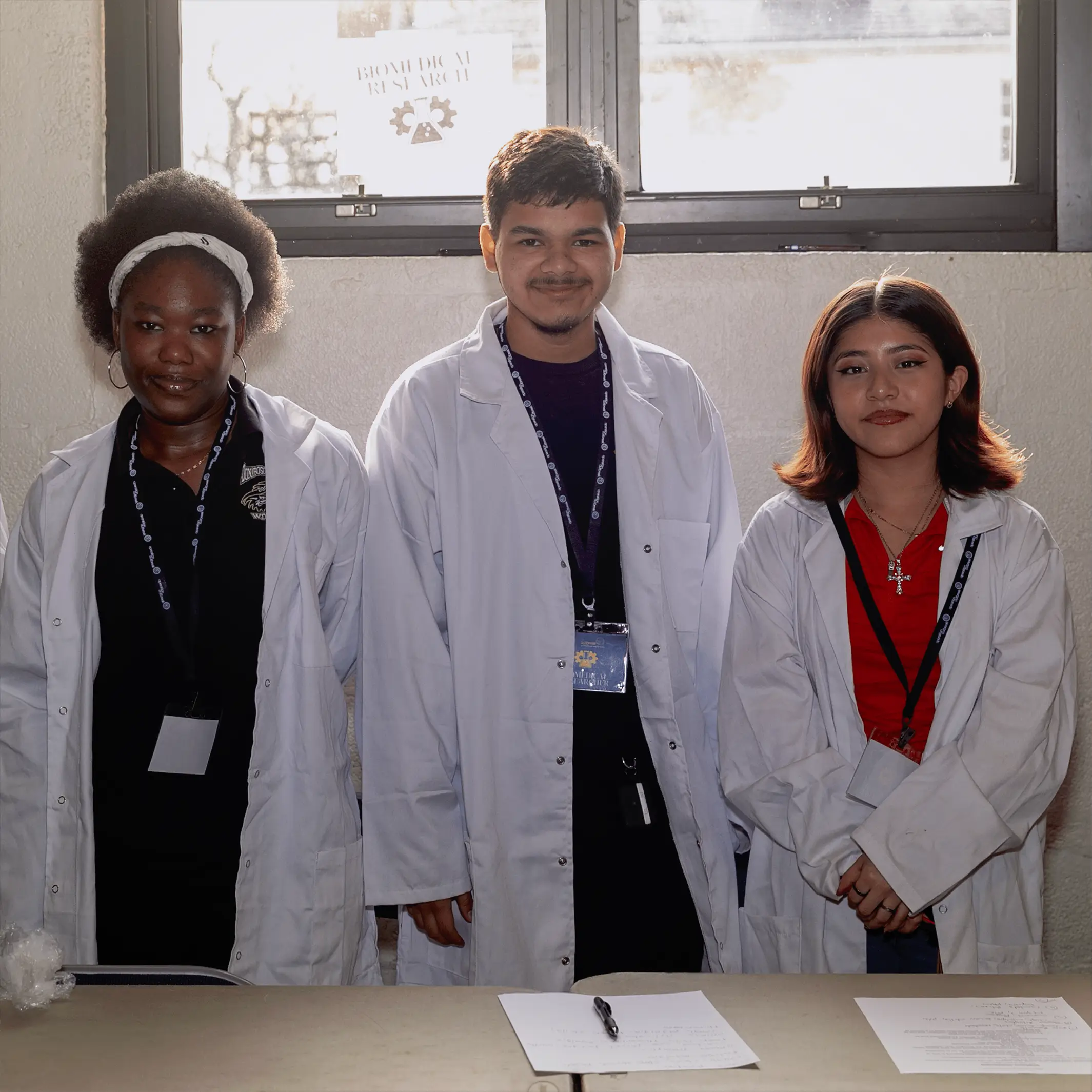 Three young adults, two women and one man, stand indoors wearing white lab coats and ID lanyards. Behind a table with papers and pens, they appear ready to support Operation Outbreak as sunlight filters through a window behind them.