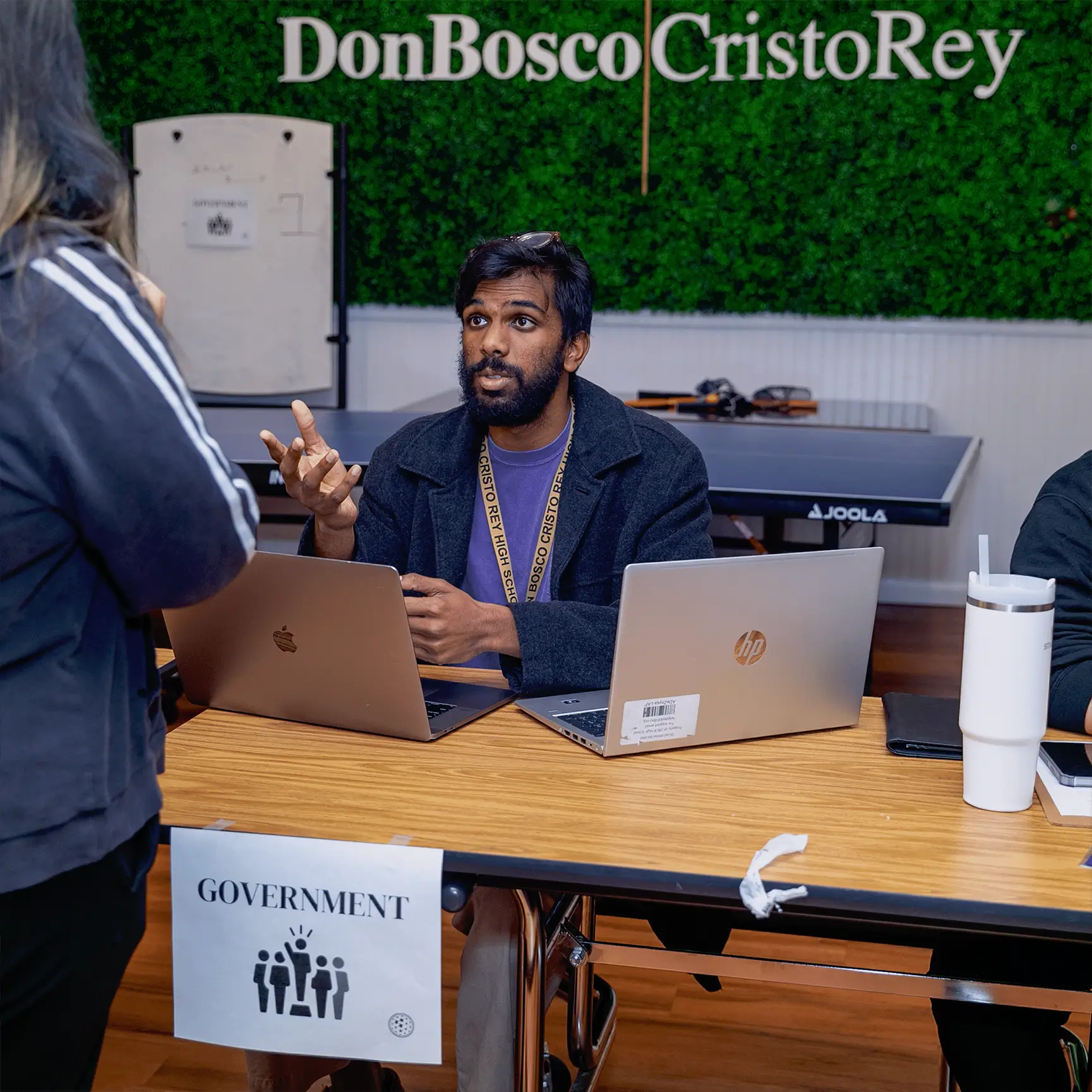A man sits at a table with two laptops, discussing Operation Outbreak with a woman standing in front of him. The table displays a "GOVERNMENT" sign, and the green wall behind them reads "Don Bosco Cristo Rey.