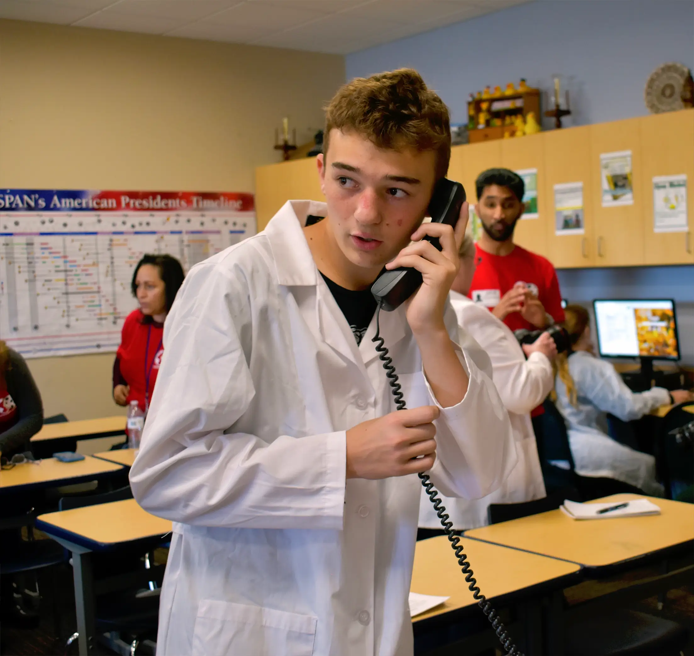 A teenage boy in a white lab coat talks on a corded phone in a classroom during Operation Outbreak, with other students and adults working in the background. Educational posters and computers are visible in the room.