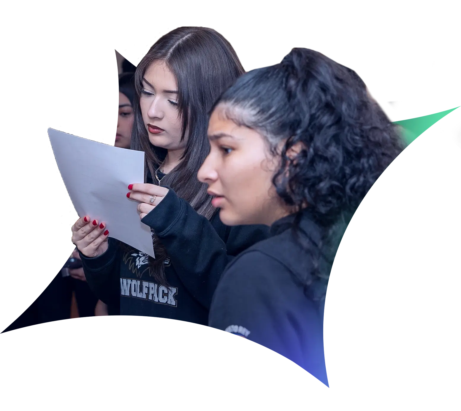 Two young women stand indoors during Operation Outbreak; one reads a sheet of paper intently while the other looks ahead, slightly out of focus. Both wear dark clothing, with other participants visible in the blurred background.