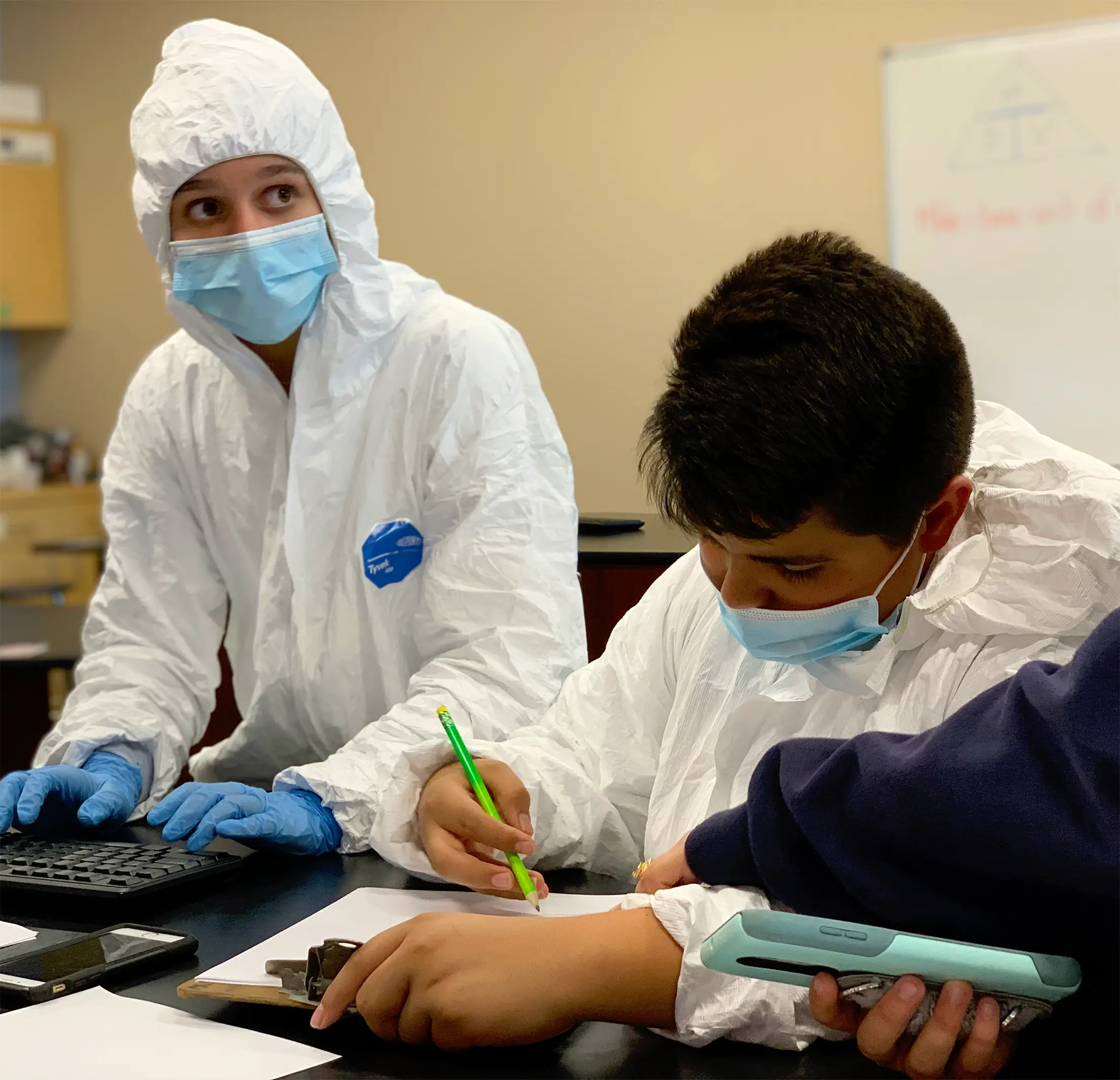 Two students in white protective suits, masks, and gloves work together at a table during Operation Outbreak. One writes with a pencil on paper while the other looks on attentively, with science equipment and a whiteboard in the background.