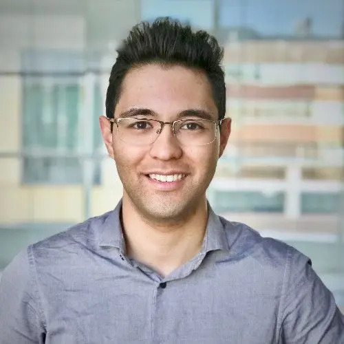 A young man wearing glasses and a light gray button-up shirt smiles at the camera. The background is blurred, showing large windows and a building outside.