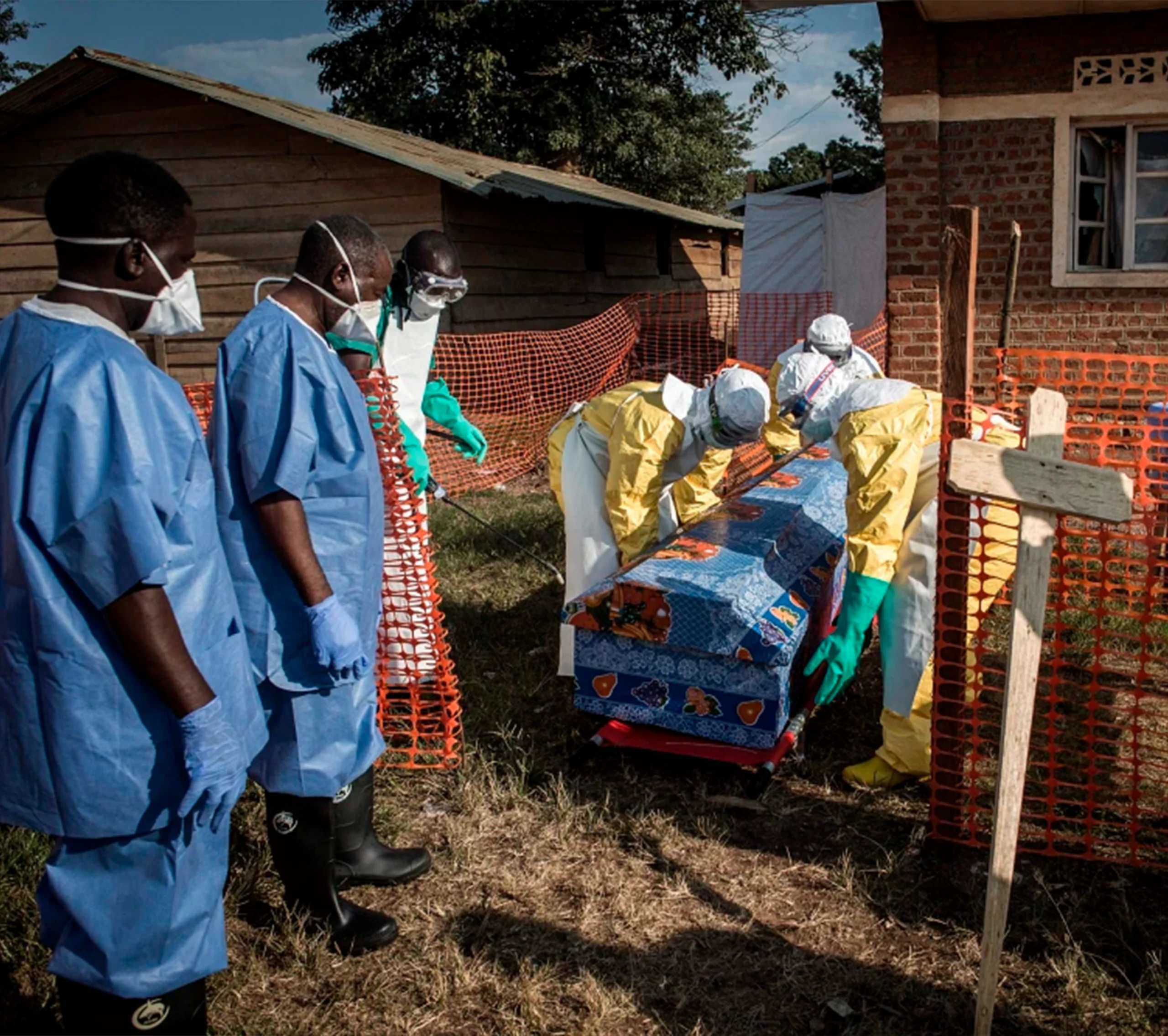 Healthcare workers in protective gear handle a covered coffin behind orange safety fencing outside a building, highlighting Operation Outbreak precautions during an infectious disease crisis.