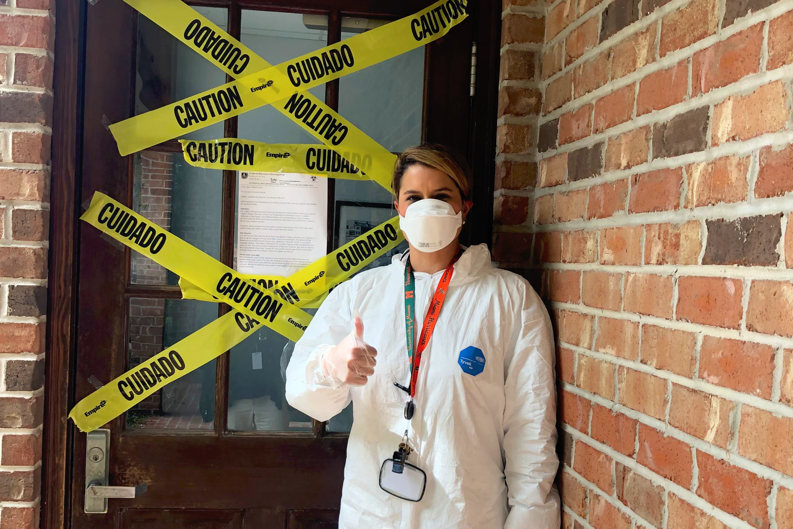 A person in protective gear and a face mask stands in front of a door covered with yellow caution tape and an Operation Outbreak notice, giving a thumbs up. The surrounding walls are brick.