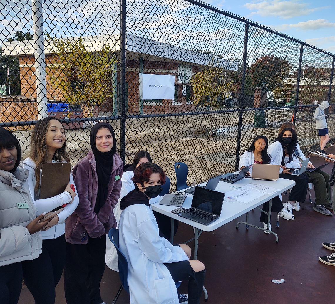 A group of people, some wearing white coats and masks, gather for Operation Outbreak around a table with laptops and papers on an outdoor basketball court near a chain-link fence. Buildings and trees are visible in the background.