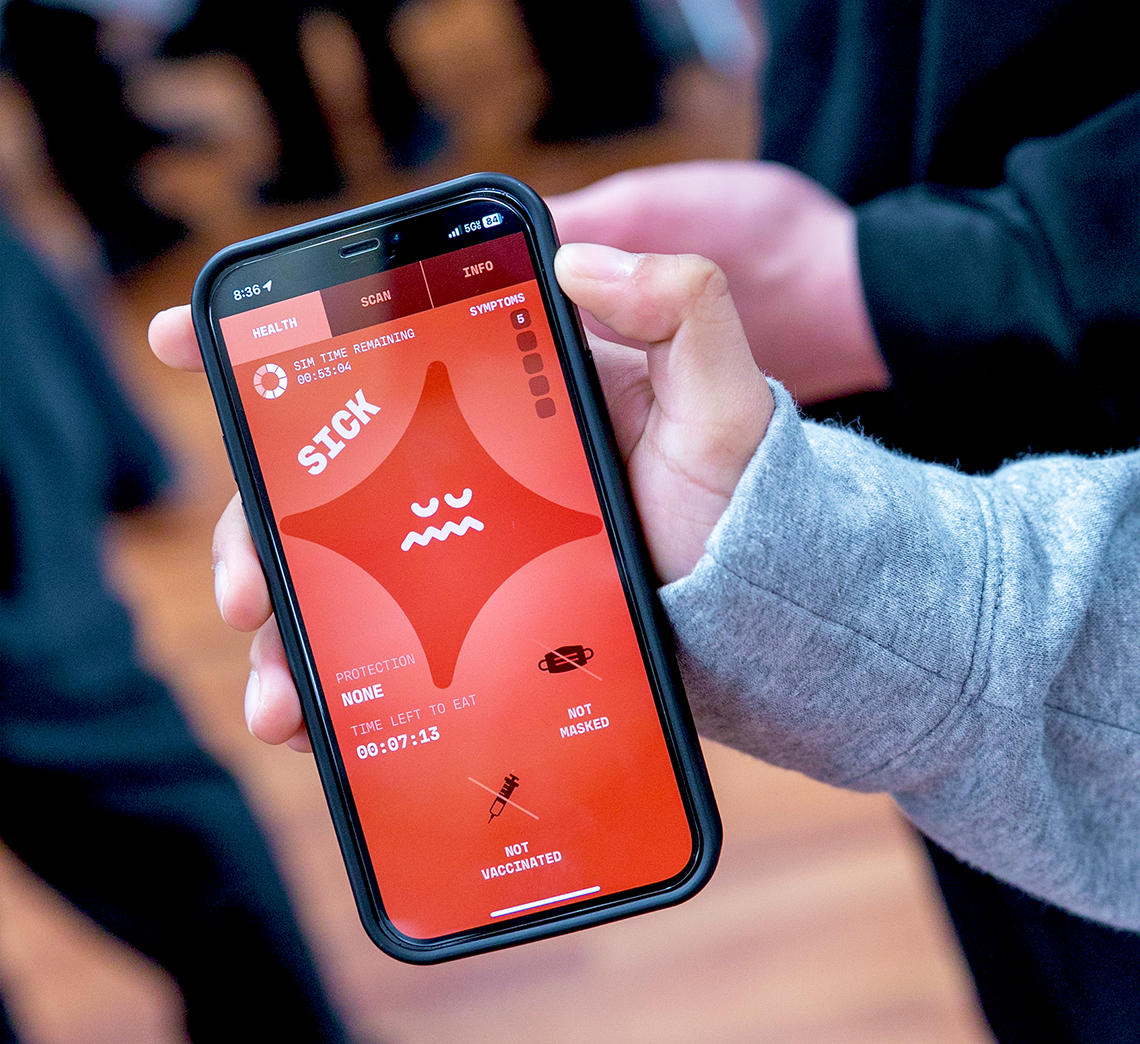 A person holds a smartphone displaying the Operation Outbreak health app with a red warning screen indicating "SICK" status, a timer, and icons for mask and vaccination status. Blurred people are visible in the background.