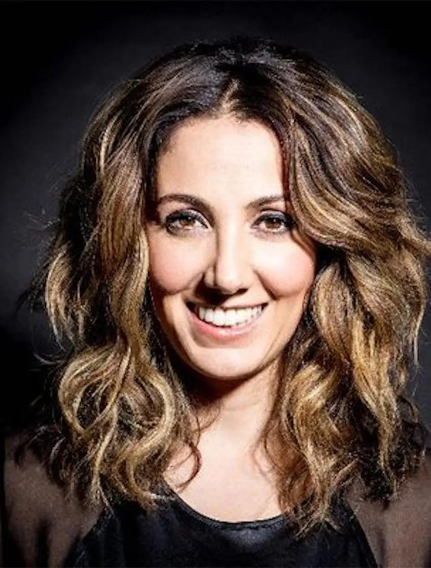 A woman with wavy, shoulder-length brown hair and highlights smiles at the camera against a dark background. She is wearing a black top and a dark jacket.