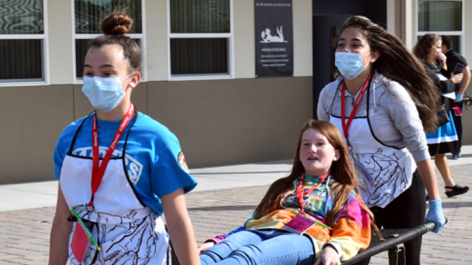 Two girls wearing masks and aprons carry another girl, who is smiling and wearing a colorful hoodie, on a stretcher outside a building during what appears to be a drill or exercise.