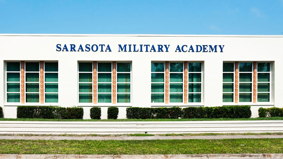 The front view of Sarasota Military Academy, a white building with large windows, shrubs, and the school name displayed in blue letters above the entrance.