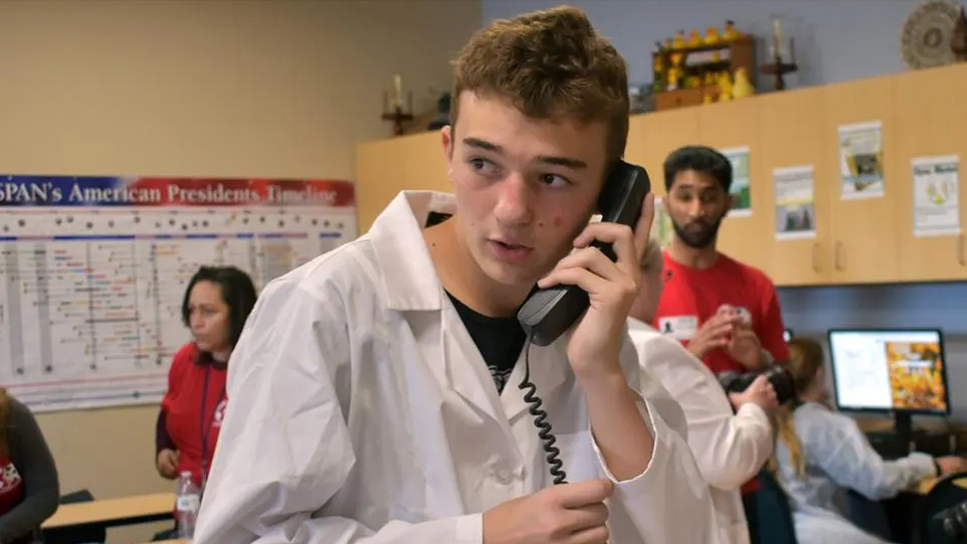 A teenage boy in a white lab coat holds a corded phone to his ear, looking to the side. Behind him, adults and children work in a classroom with science posters and computers.