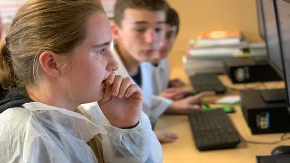Three students wearing lab coats sit at a desk, focusing intently on computer screens. The foreground shows a girl with her hand on her chin, while two boys are blurred in the background.