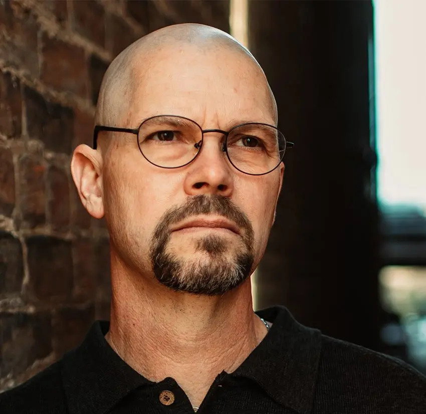 A bald man with glasses and a goatee stands against a brick wall, wearing a black collared shirt and looking thoughtfully into the distance.