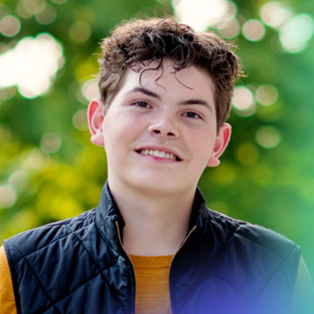 A young person with short curly hair smiles outdoors, wearing a black quilted vest over a mustard yellow sweater, with a blurred green background of trees.
