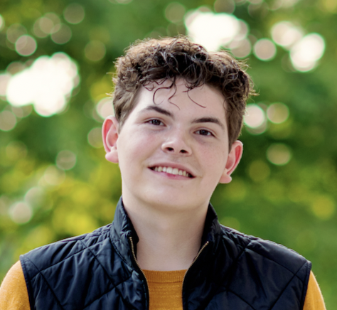 A young person with short curly hair, wearing a mustard yellow long-sleeve shirt and a dark quilted vest, stands outdoors between two white columns, with green blurred trees in the background.