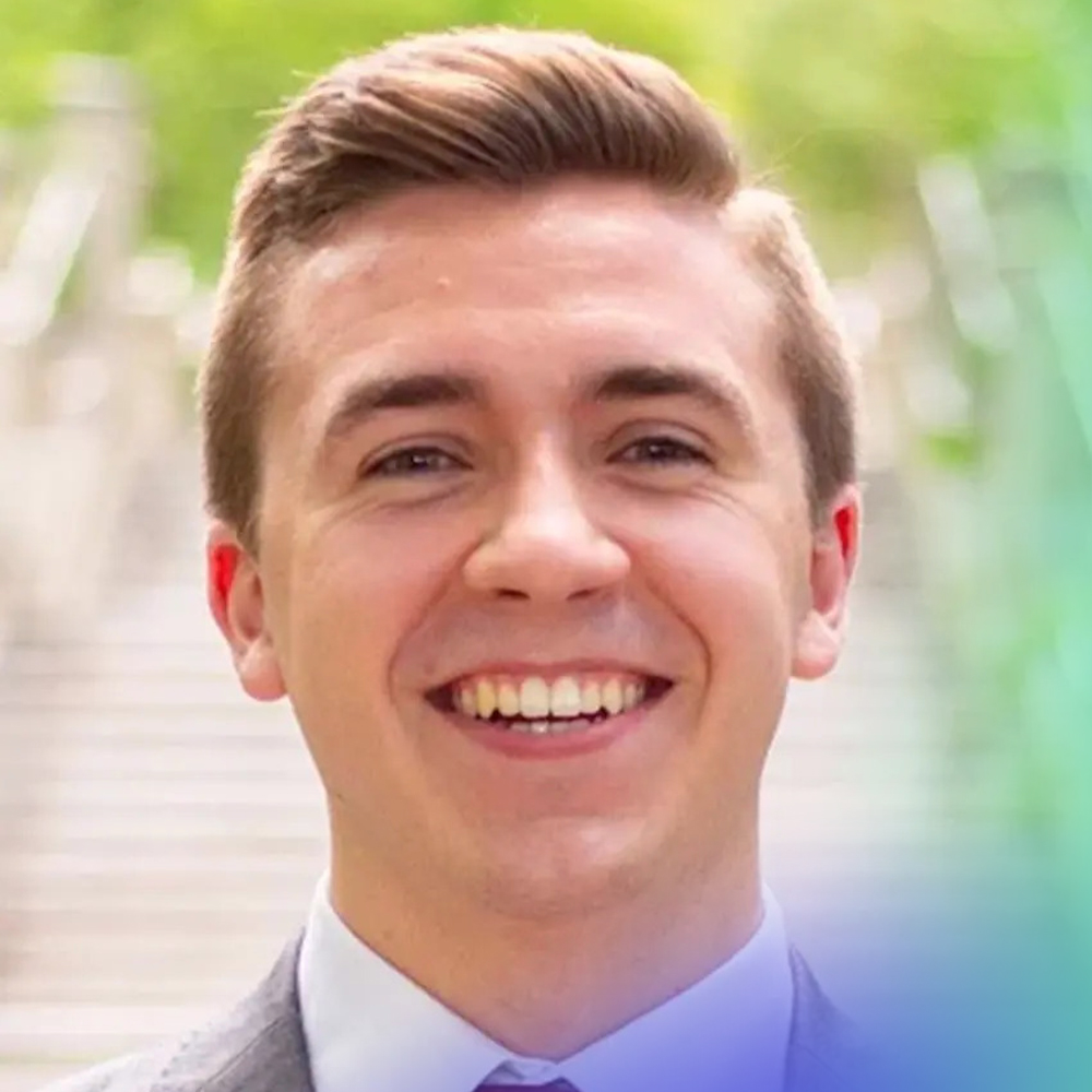 A young man in a suit smiles broadly outdoors, with blurred green foliage and steps in the background.