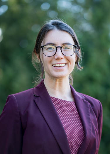 A woman with glasses and brown hair tied back, wearing a maroon blazer over a patterned top, smiles outdoors with greenery blurred in the background.