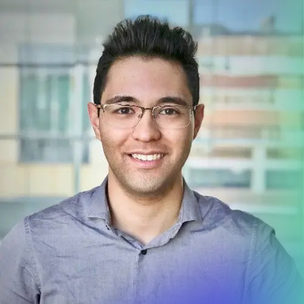 A young man with short dark hair and glasses smiles at the camera, wearing a light gray button-up shirt. The background shows a blurred indoor setting with large windows and some outdoor light.