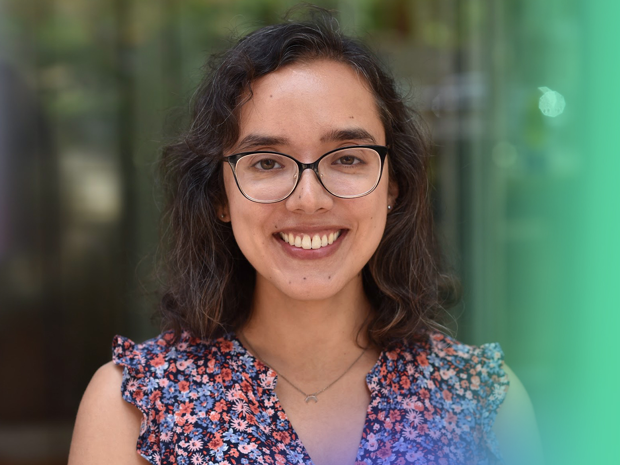 A woman with curly brown hair, glasses, and a floral blouse smiles at the camera. The background is blurred with shades of green and brown.