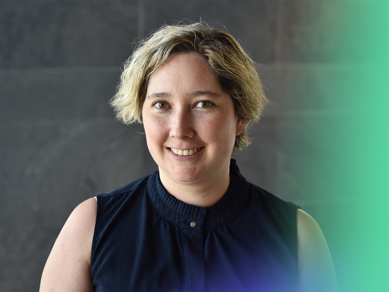 A person with short, wavy blond hair and a sleeveless dark top smiles at the camera, standing in front of a dark, blurred background with green and blue light streaks on the right side.