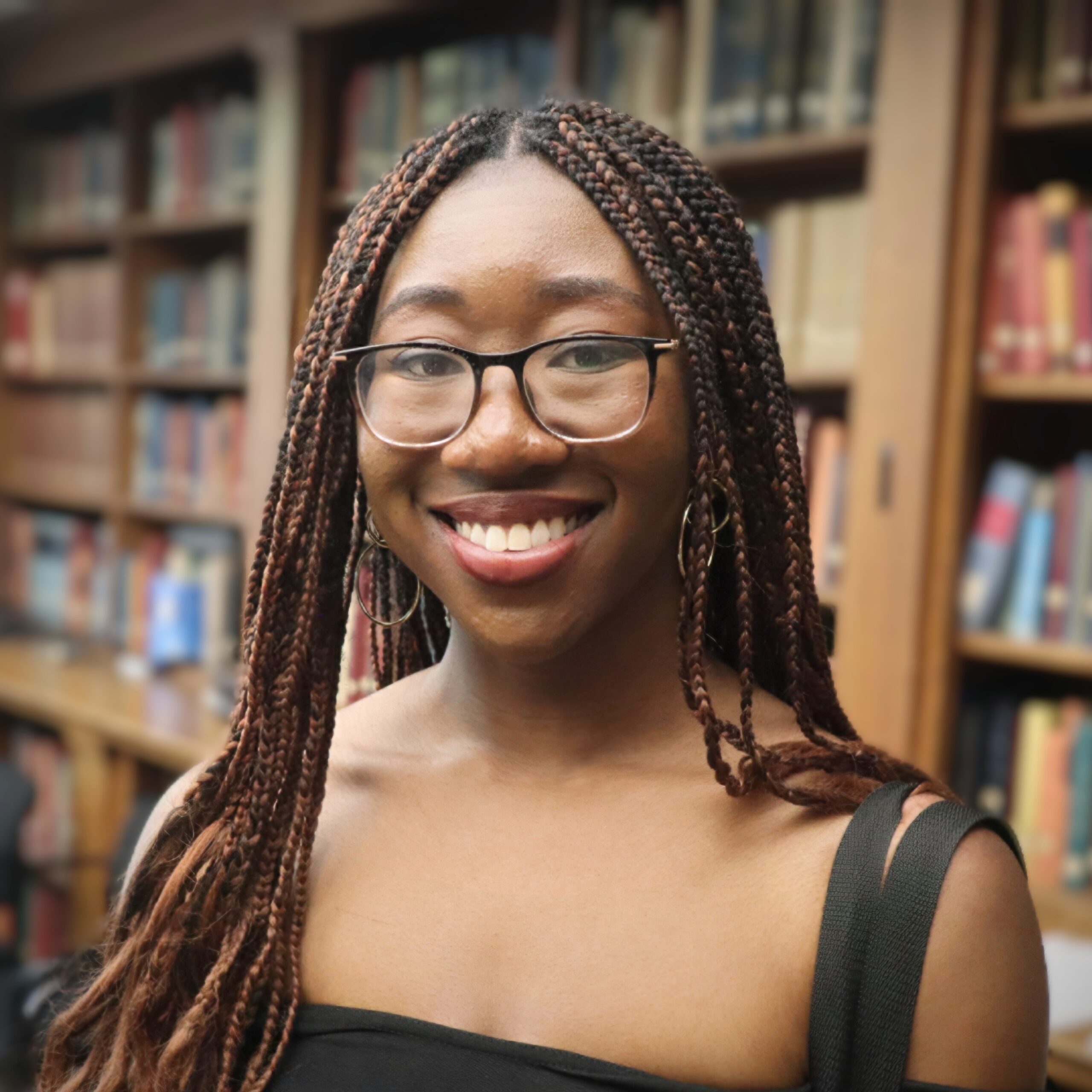 A young woman with glasses and braided hair smiles in front of bookshelves filled with books. She is wearing hoop earrings and a black off-shoulder top.