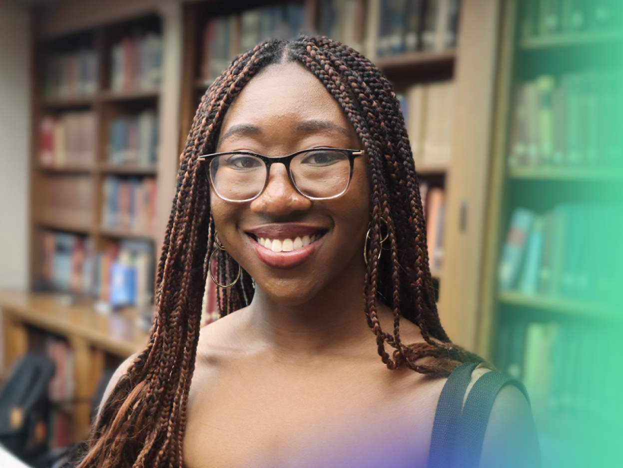 A woman with braided hair and glasses smiles in a library, standing in front of bookshelves filled with books. The foreground has blue and green blurred areas.