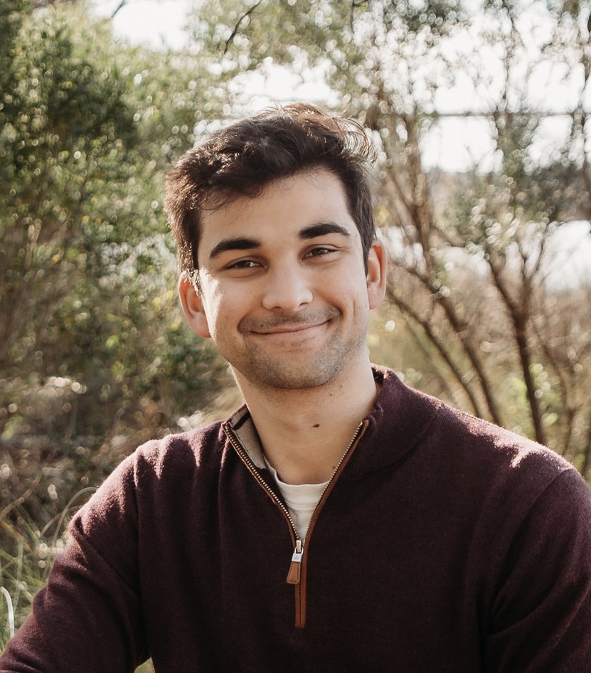 A young man with dark hair and a slight smile sits outdoors, wearing a maroon sweater. Sunlight filters through green trees and foliage in the background.