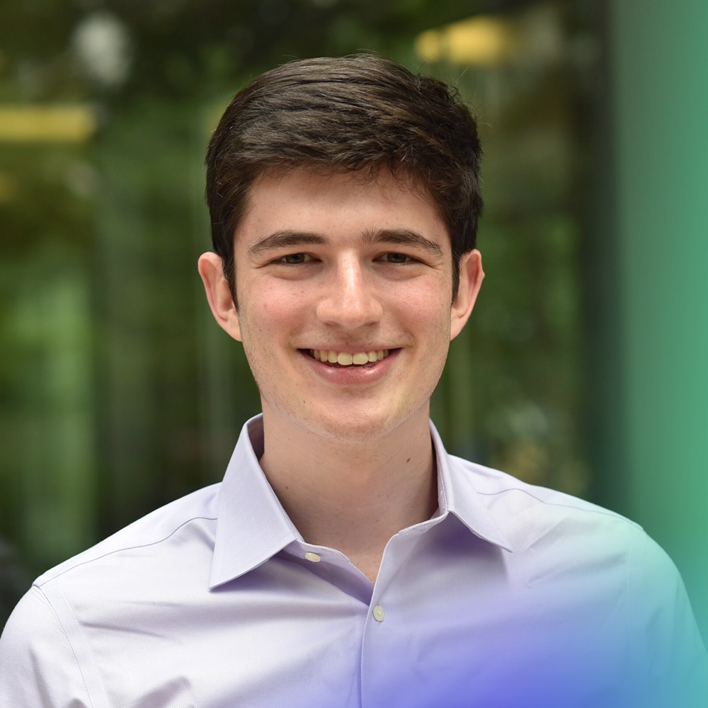 A young man with short brown hair, wearing a light purple button-up shirt, smiles at the camera. The background is blurred greenery with hints of yellow and blue near the edges of the image.