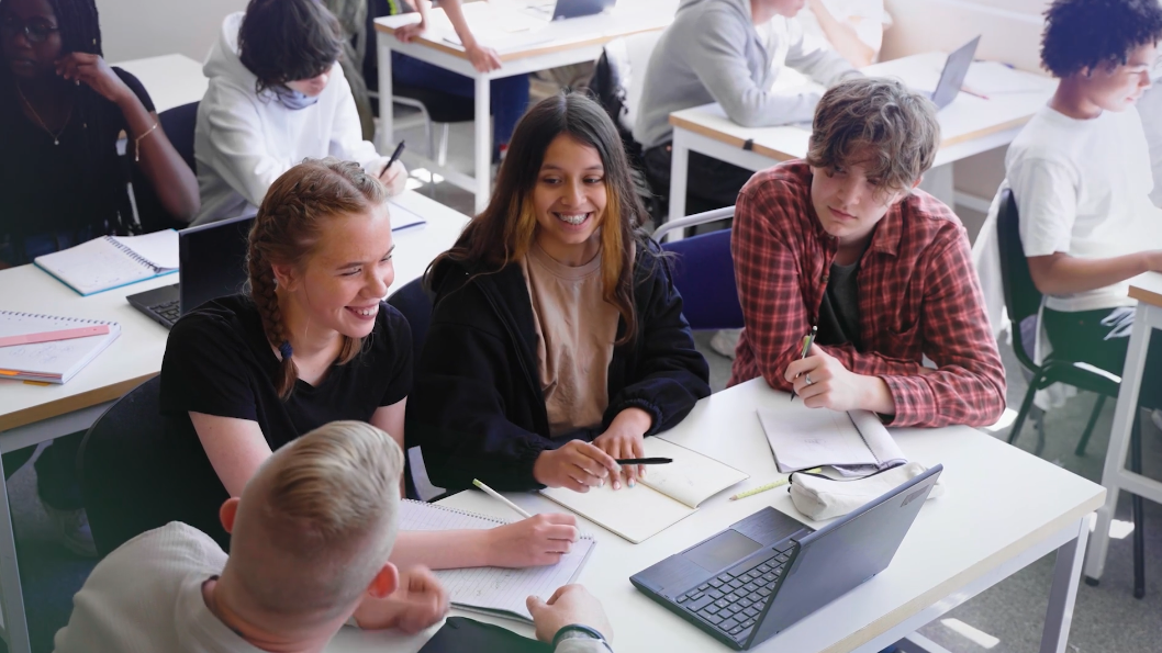 A group of four students sit around a table, smiling and talking, with notebooks, papers, and a laptop open to Operation Outbreak. Other students work at tables in the background in a bright classroom.