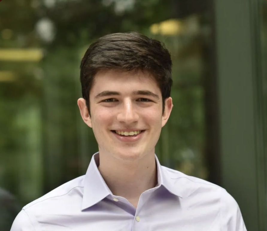 A young man with short dark hair, wearing a light purple collared shirt, smiles while standing in front of a blurred green outdoor background.