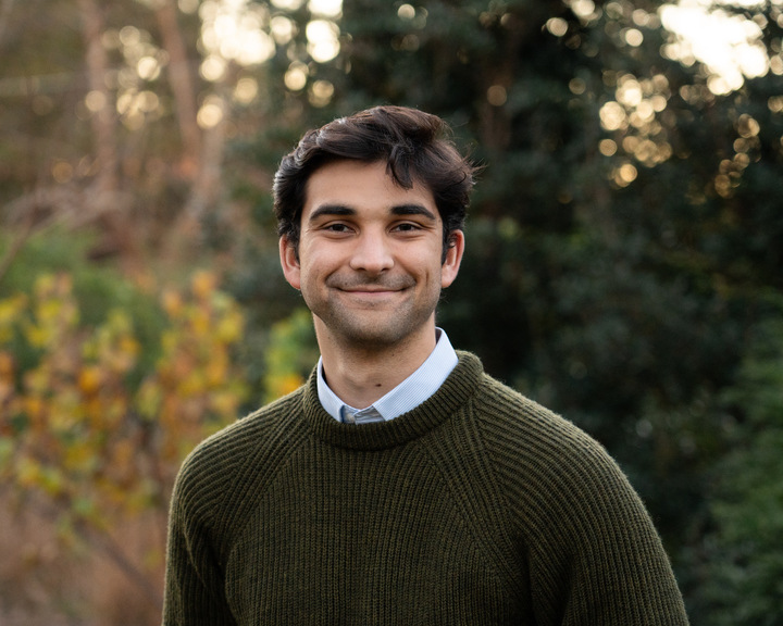 A young man with dark hair smiles while standing outdoors in front of trees, wearing a green knit sweater over a collared shirt. The background is softly blurred with autumn foliage.