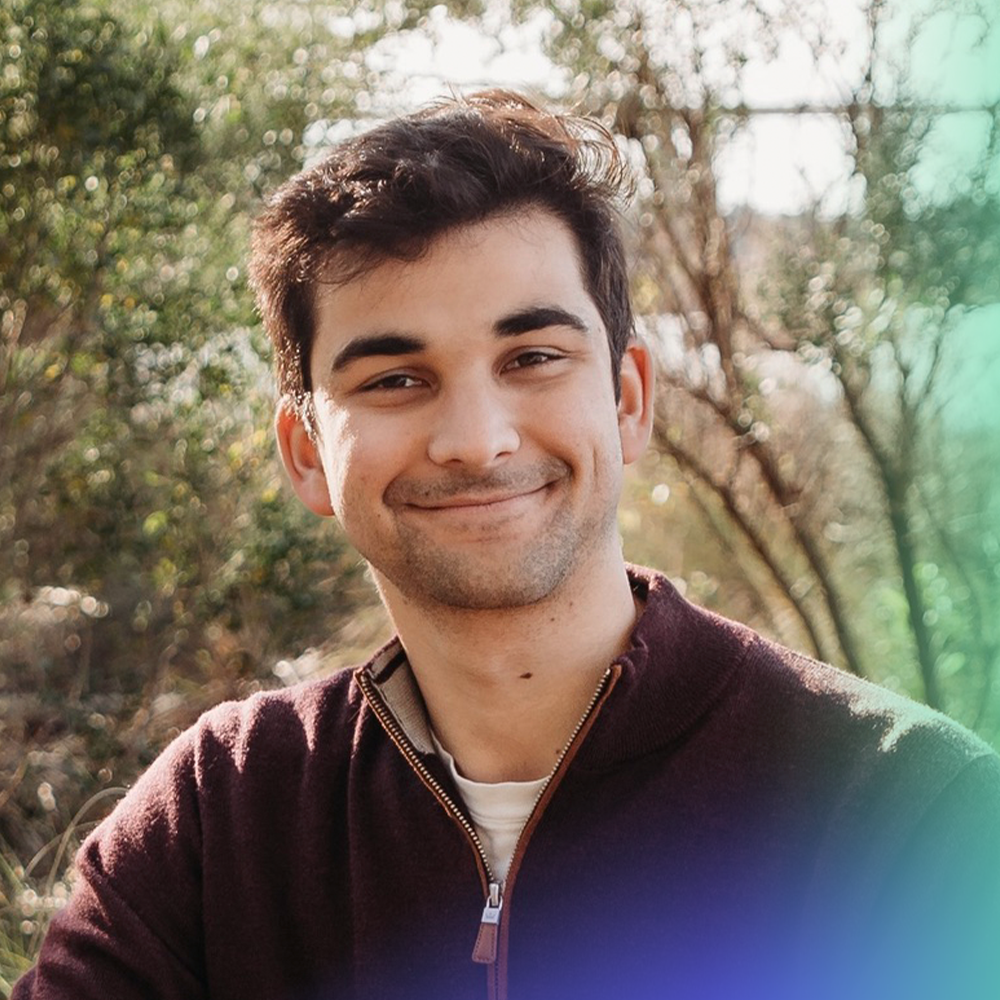 A young man with dark hair and a trimmed beard smiles at the camera while sitting outdoors, surrounded by greenery and sunlight, wearing a maroon zip-up sweater over a light shirt.