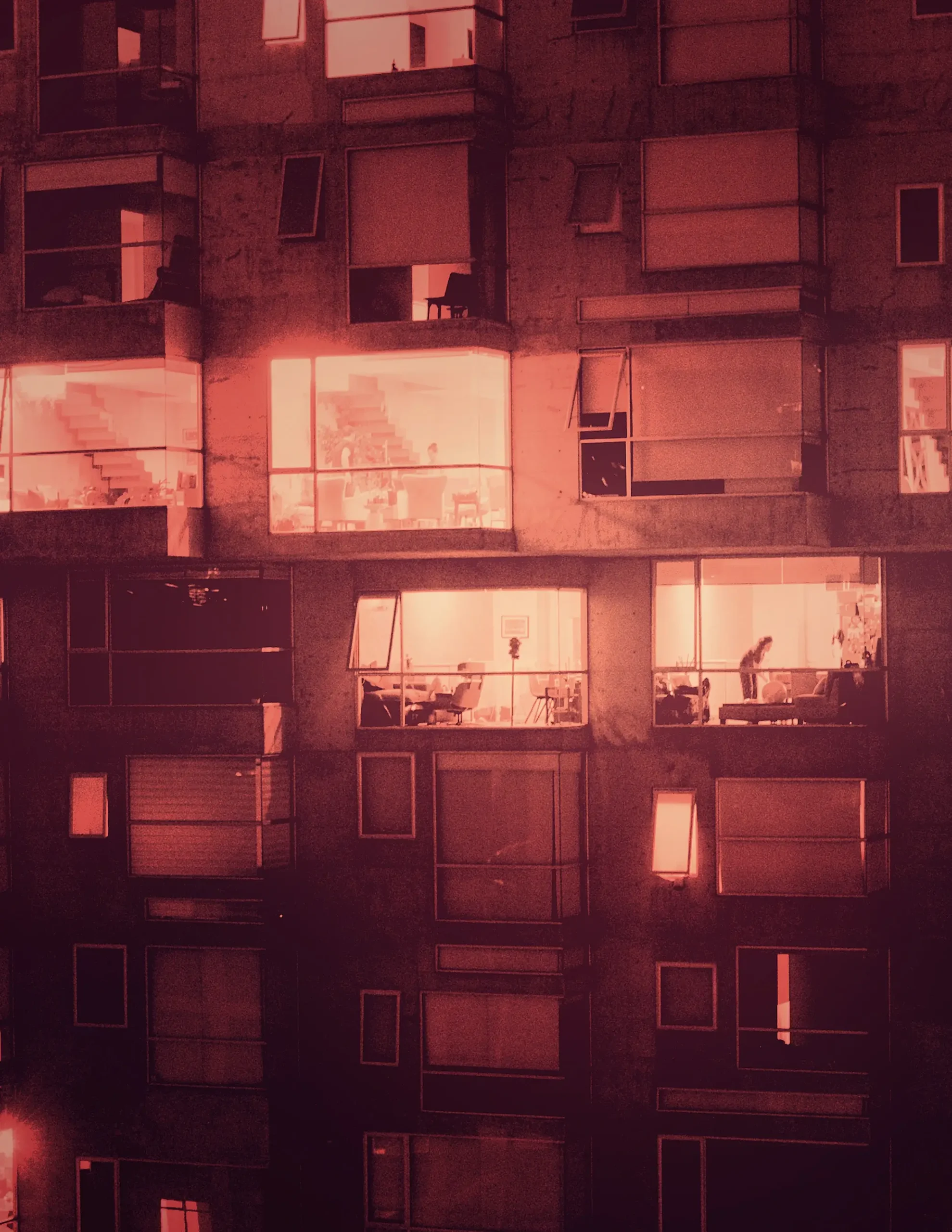 A nighttime view of an apartment building with large windows glowing orange from interior lights, showing glimpses of people and furniture inside several units during Operation Outbreak. Some windows have blinds partially drawn.
