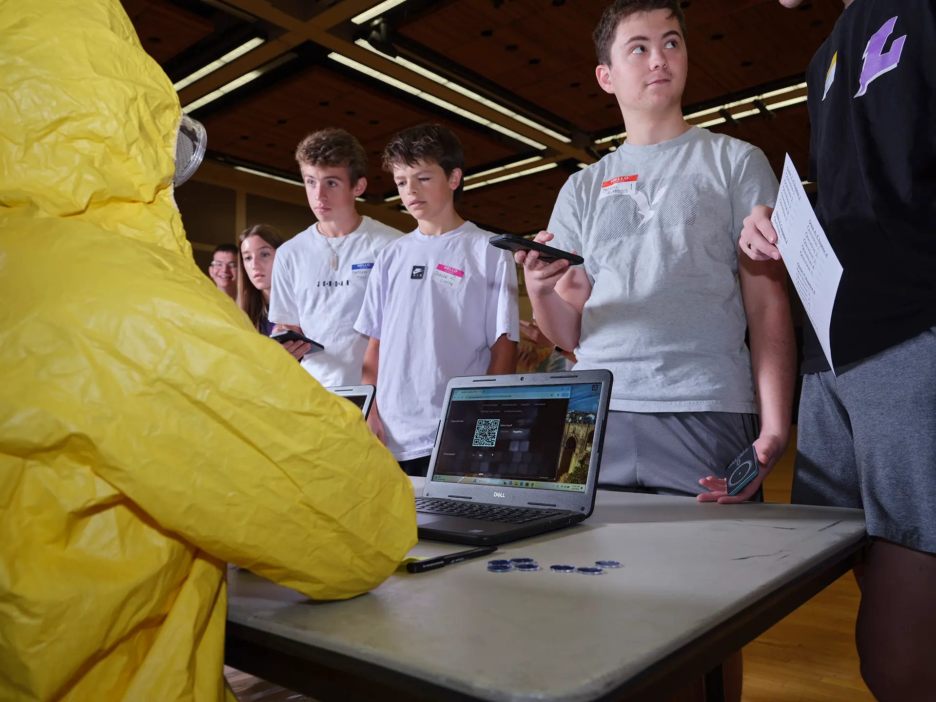A person in a yellow hazmat suit sits at a table with a laptop, while a group of teenagers stand in line, holding papers or phones, waiting to interact indoors.