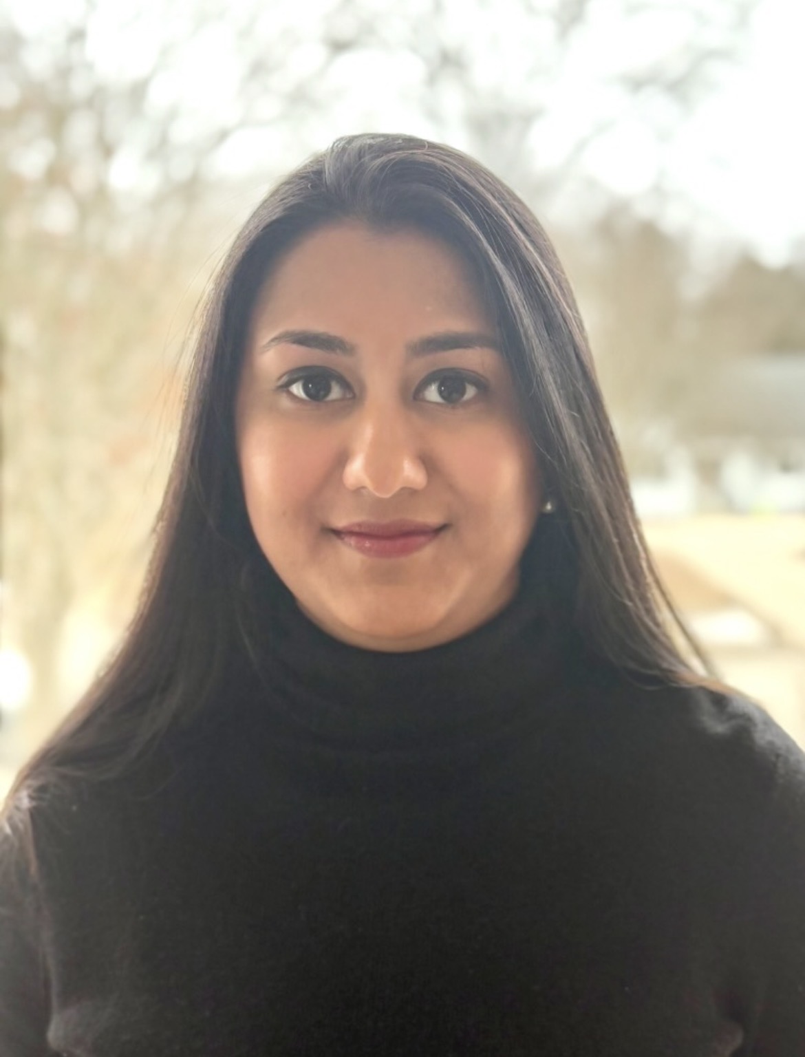 A woman with long, dark hair and a black turtleneck sweater looks at the camera, smiling softly. The background is softly blurred, showing a bright outdoor scene with trees and light.