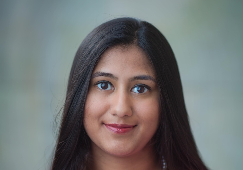 A young woman with long, straight dark hair and brown eyes smiles gently at the camera. She wears light makeup and a pearl necklace, with a soft, blurred background.